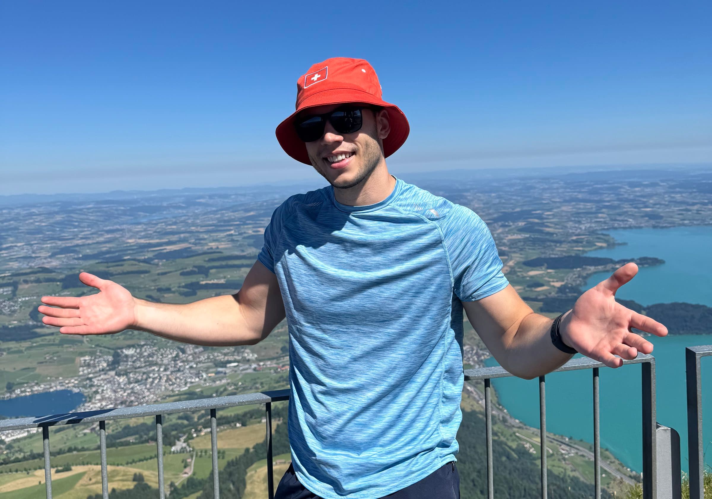 Me on a summit above a lake, arms wide open, in a red Swiss bucket hat.